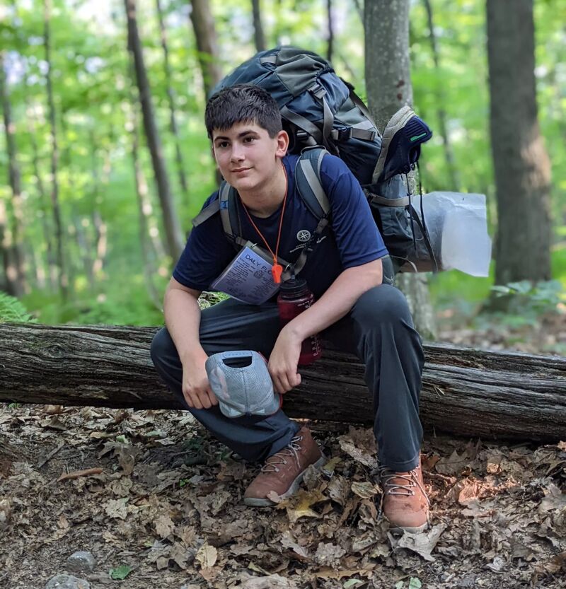 A young man is sitting on a fallen log in a forest. He is wearing a dark blue t-shirt, dark pants, and hiking boots. He has a large backpack behind him. The forest is green and lush, with tall trees and fallen leaves on the ground. The lighting suggests it is daytime.